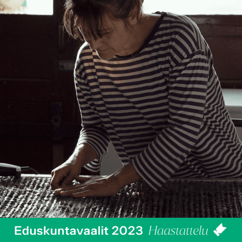 Kuvassa kuvataiteilija Hanna Vihriälä seisoo pöydän ääressä, kumartuneena työskentelemään teoksen äärellä niin, etteivät taiteilijan kasvot näy. Kuvan alareunassa on erillisessä väripalkissa teksti: "Eduskuntavaalit 2023 / Haastattelu".