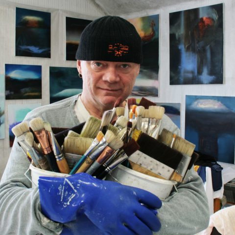 Taiteilija Ismo Hyvärisen omakuva, jossa hän pitelee sylissään pensseleitä täynnä olevia purkkeja. Taustalla seinillä näkyy hänen teoksiaan.