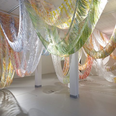 Kuva gallerian näyttelytilasta, jonne on ripustettu katosta roikkumaan lomittain erivärisiä, käytöstä poistettuja lakanoita, joita taiteilija Johanna Pöykkö on työstänyt leikkaamalla, maalaamalla ja ompelemalla.