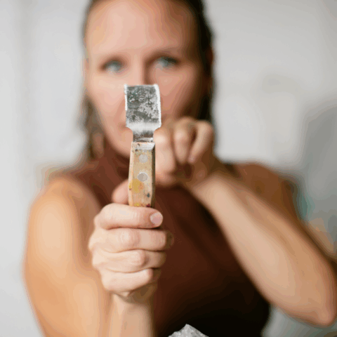 Valokuvassa kuvanveistäjä pitelee toisessa kädessään taltan kaltaista työkaluaan, johon kamera tarkentaa ja taempana oleva taiteilija erottuu sumeampana.